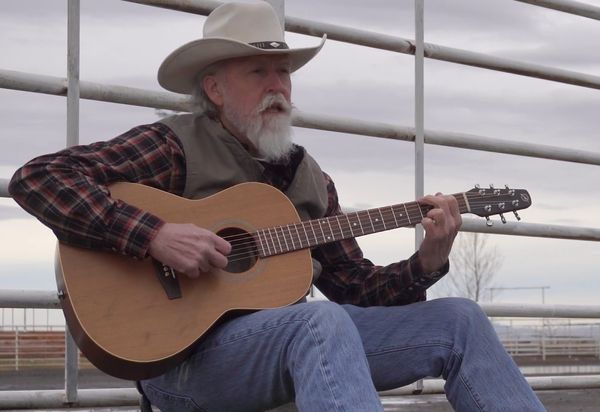 Musician in a cowboy hat playing guitar beside a corral fence